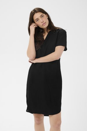 Kvinde med langt brunt hår iført en kortærmet sort kjole, der står med den ene arm over kors og den anden hånd rører ved sit hår.