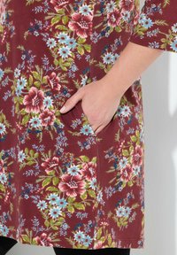 Blumen-Tunika mit maroonfarbener Basis, die große rote und blaue Blumen sowie grüne Blätter zeigt, und seitlichen Taschen. Aus weichem Stoff gefertigt, mit kurzen Ärmeln bis zum Ellenbogen.