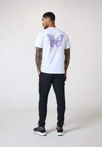 Hvid T-shirt med et lilla sommerfugl grafik og design af dryppende maling på ryggen. Kombineret med sorte cargo-bukser og sneakers.