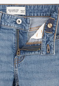 Visoke ravne traperice svijetloplave boje. Imaju zlatni patentni zatvarač, mesingano dugme za zatvaranje i kontrastni narančasti šav. Oznaka je vidljiva.