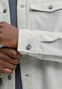 Camisa de franela gris de lana con puños abotonados, que presenta una superficie texturizada y botones de color gris oscuro. Incluye un detalle de bolsillo en el pecho.
