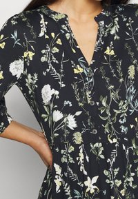 Vestido negro con un patrón floral en verde, blanco y amarillo, con escote en V y tres botones, y mangas de 3/4; brazo doblado, mano resting on la cadera.