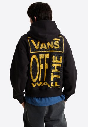 Henkilö, jolla on päällään musta huppari, jossa suuri keltainen Vans Off The Wall -logo selässä, sininen paita alla ja tummat housut.