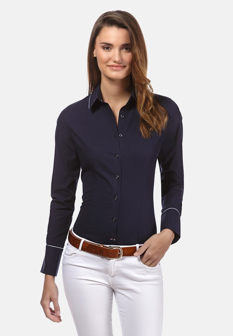 Camicia blu navy con maniche lunghe, bordi bianchi su polsini e colletto, abbinata a pantaloni bianchi e una cintura marrone. Tessuto a trama liscia.