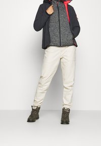 Tenue de randonnée comprenant une veste noire à capuche, doublée en polaire grise, un pantalon beige clair et des bottes marron à lacets avec une finition texturée.