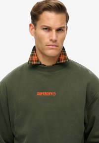 Olijfgroene sweatshirt met het "SUPERDRY" logo in oranje, gelaagd over een zwart en oranje geruit overhemd met kraag. Zachte stof en een relaxte pasvorm.