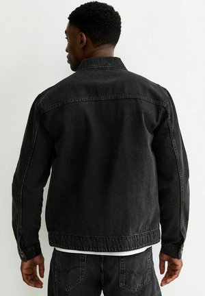 Man in een zwarte spijkerjack en bijpassende zwarte spijkerbroek, met de rug naar de camera, tegen een effen witte achtergrond.