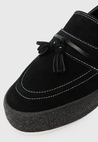 Czarne mokasyny zamszowe z kontrastującym białym przeszyciem, ozdobione detalem frędzli i teksturowaną czarną gumową podeszwą.