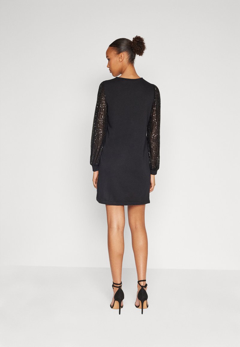 Robe noire avec de longues manches pailletées, coupe droite, encolure ronde et longueur au-dessus du genou, accompagnée de sandales à talons noires.