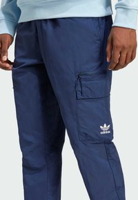 Granatowe spodnie cargo wykonane z lekkiego materiału, z boczną kieszenią na zamek błyskawiczny i haftowanym białym logo Adidas.