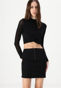 Čierny, priliehavý crop top s dlhými rukávmi a prekrúteným predným dizajnom, skombinovaný s čiernou džínsovou sukňou s vysokým pásom a zipsom.