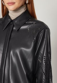 Femme portant une chemise en cuir noire avec des motifs floraux découpés au laser sur l'épaule et la manche, associée à un col roulé beige et des boucles d'oreilles anneaux argentées.