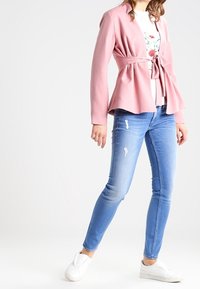 Ljusrosa wrap blazer med knytmidja, kombinerad med en vit blommig skjorta och ljusblå slitna skinny jeans. Vita slip-on sneakers.