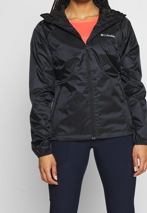 Femme portant une veste noire Columbia zippée avec capuche, sur un t-shirt corail, associée à des leggings foncés, debout devant un fond neutre.