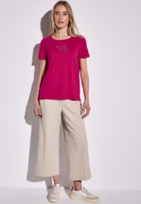 T-shirt fuchsia avec l'imprimé "NEVER DOUBT yourself", associé à un pantalon large beige et des baskets beiges avec des accents roses.