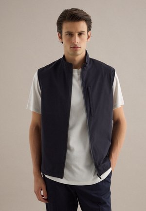 Uomo che indossa un gilet smanicato blu navy con cerniera sopra una maglietta bianca a maniche corte, in piedi davanti a uno sfondo beige uniforme.