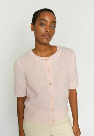 Frau mit kurzen Haaren, die eine hellrosa, kurzärmlige, knopfbesetzte Strickjacke und beige Hosen trägt, steht vor einem schlichten weißen Hintergrund.