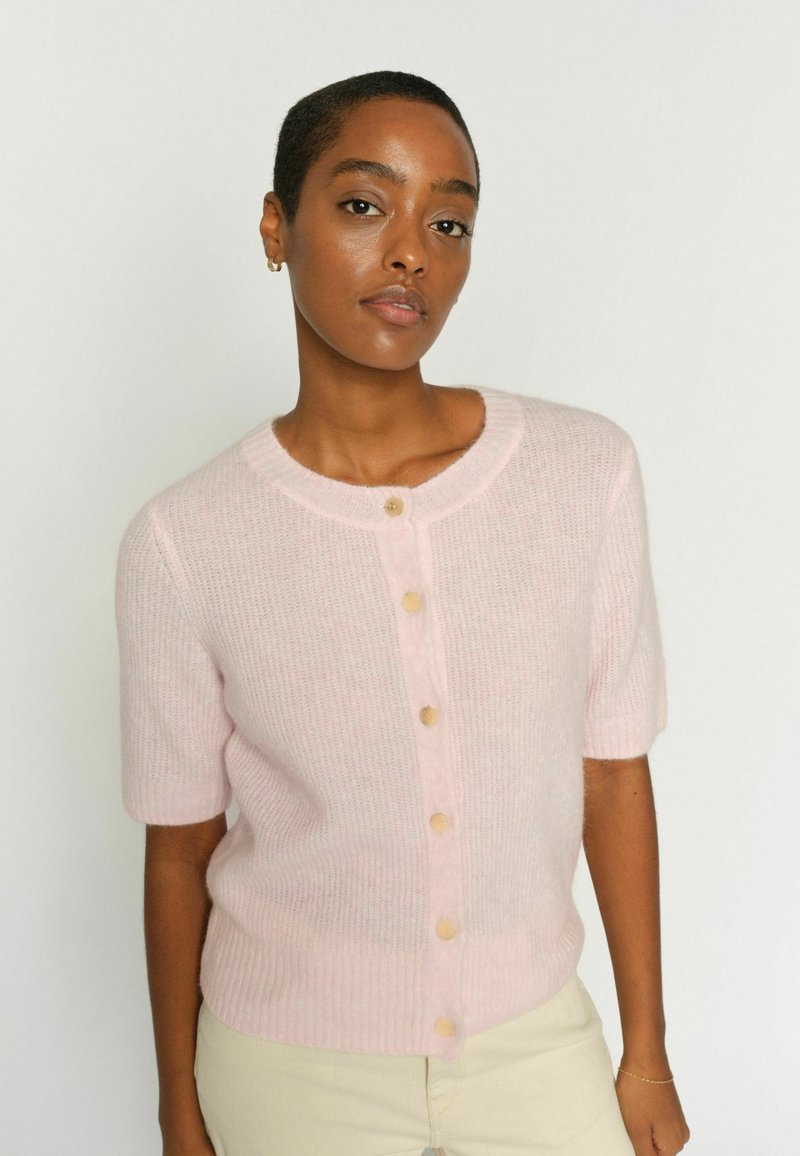 Sieviete ar īsiem matiem, ģērbusies gaiši rozā, īss piedurkņu, poga aiztaisīta kardigānā un bēšās biksēs, stāv uz vienkārša baltā fona.