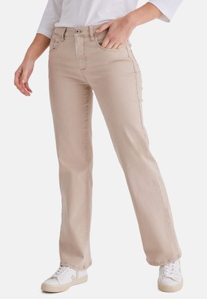 Frau trägt beige Jeans mit geradem Bein, weiße Sneaker und ein weißes Hemd, mit einer Hand in der Tasche vor einem schlichten Hintergrund.