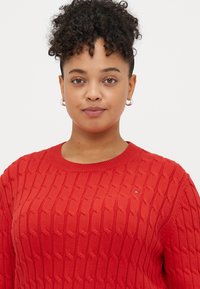 Rød strikket genser med kabelmønster, ribbestrikket halslinning og liten logo aksent på brystet. Myk tekstur og avslappet passform.