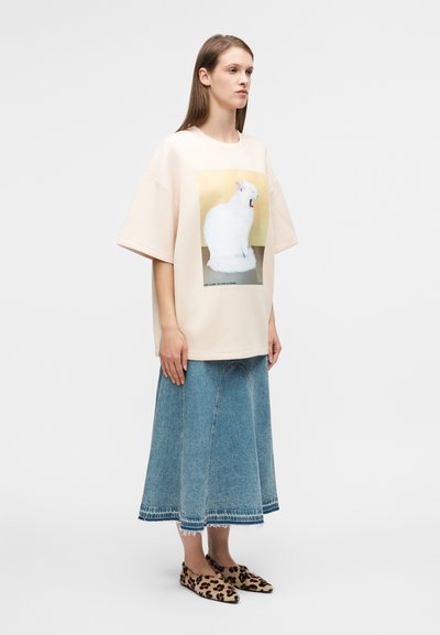 Μπεζ oversized μπλουζάκι με γραφικό γάτας, συνδυασμένο με μπλε τζιν φούστα και παπούτσια slip-on με λεοπάρ σχέδιο. Λείο ύφασμα και άνετη εφαρμογή.