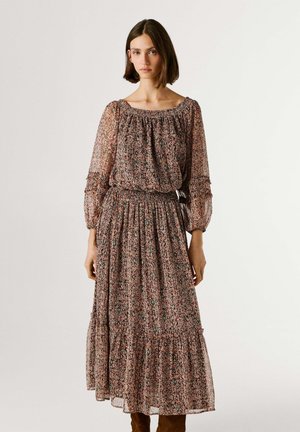 Junge Frau in einem langen, gemusterten Kleid mit durchsichtigen, aufgebauschten Ärmeln und tailliertem Bund, die vor einem einfarbigen hellen Hintergrund steht.