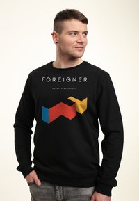Felpa nera con la parola "FOREIGNER" sopra un design geometrico colorato in rosso, blu e giallo. Il tessuto sembra morbido e presenta un collo tondo.