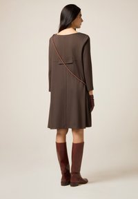 Bruine, knielange jurk met een rechte snit, lange mouwen en een strikdetail aan de achterkant. Gecombineerd met bruine kniehoge laarzen en een tas met een dunne riem.