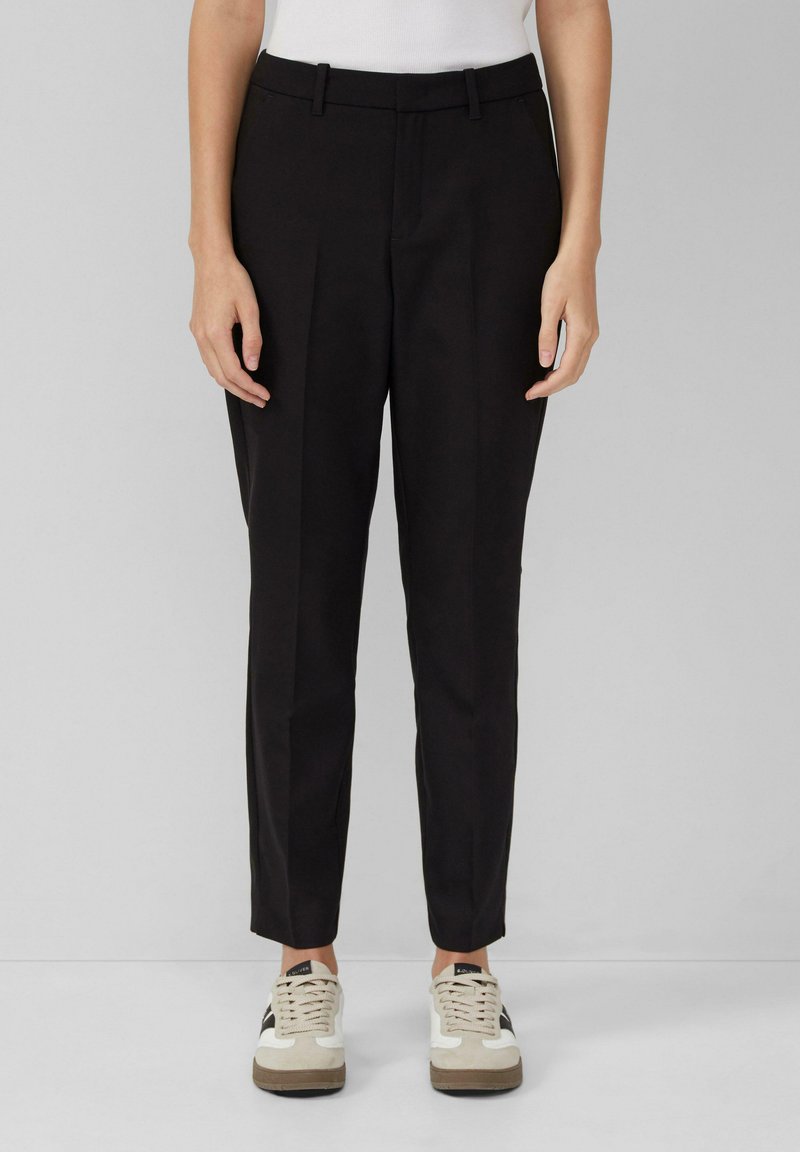 Pantalons noirs ajustés avec une taille plate, des jambes fuselées et des passants de ceinture, associés à des baskets beige et noires pour un look décontracté.