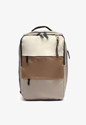 Sac à dos rectangulaire tricolore avec des sections beige, blanche et marron, plusieurs compartiments zippés, des tirettes de fermeture éclair noires, et un porte-clés tressé attaché.