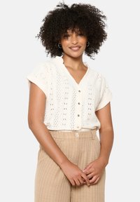 Nertinė trumpomis rankovėmis kreminės spalvos bluzonė su V formos iškirpte, sagomis priekyje ir atviru raštu. Derinama su smėlio spalvos dryžuotomis, aukšta juosena kelnių.