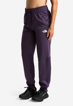 Person trägt violette The North Face Jogginghose und schwarze Sneakers, steht mit sichtbarem Tattoo am rechten Arm.