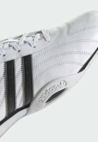 Biele kožené tenisky s tromi čiernymi pruhmi, textúrovanými detailmi a gumovou podrážkou s výrazným logom a zaobleným dizajnom špičky.