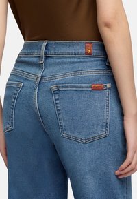 Personne portant un jean en denim bleu avec des poches arrière et des étiquettes rouges, associé à un top marron ajusté, vue de la taille vers le bas.
