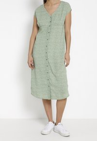Robe verte à boutons avec un décolleté en V, des manches courtes et un motif floral. Fabriquée en tissu léger, elle tombe au milieu du mollet. Portée avec des baskets blanches.