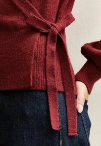 Bordowy sweter dzianinowy z wiązanym paskiem w talii, z teksturowanym ściągaczem u dołu, w połączeniu z ciemnymi dżinsami. Miękka tkanina.