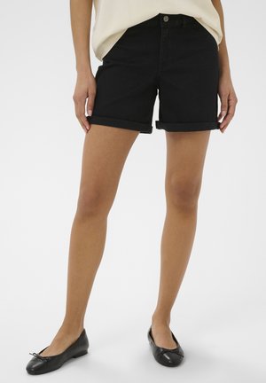 Woman wearing black rolled-hem shorts, cream top, and black ballet flats, standing against a plain white background.