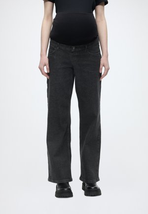 Raskaana oleva henkilön päällä mustat äitiysfarkut joustavalla vyötäröllä ja mustat paksupohjaiset saappaat, seisoo yksiväristä taustaa vasten.