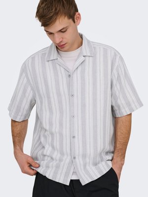 Bărbat tânăr care poartă o cămașă cu mâneci scurte, gri și alb cu dungi verticale, nasturi, peste un tricou alb, cu o mână în buzunar și capul înclinat în jos.