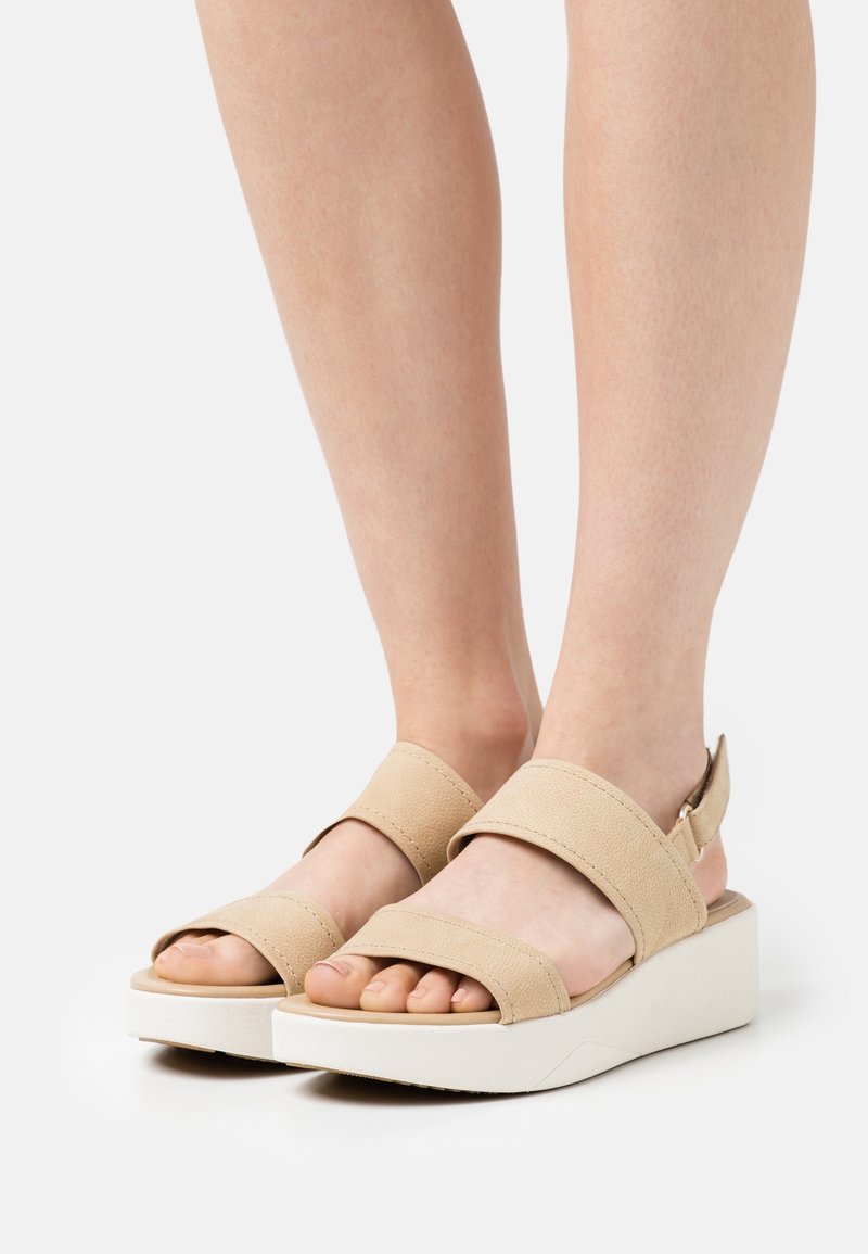Pieds portant des sandales beige à bout ouvert avec deux larges sangles et des semelles compensées blanches sur un fond clair.