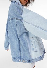Světle modrá džínsová bunda se strukturovaným designem, kombinující různé odstíny, velké kapsy a knoflíkové zapínání, které nabízí uvolněný střih.
