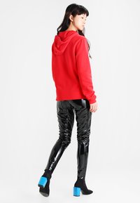 Piros kapucnis pulóver laza fazonban, fényes fekete leggings-szel és kék sarkú csizmával párosítva. Háttér sima fehér.