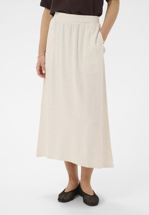 Kobieta nosząca beżową spódnicę midi z elastycznym paskiem w talii, czarne plecione baleriny oraz ciemną bluzkę, z rękami opuszczonymi wzdłuż ciała.