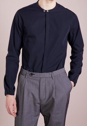 Mann trägt ein marineblaues Hemd mit Knöpfen am Kragen, in graue karierte Hose gesteckt, mit einer Hand in der Tasche vor einfarbigem Hintergrund.