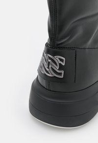 Zbliżenie obcasa czarnego skórzanego buta z srebrnym, haftowanym logo dekoracyjnym i grubą czarną gumową podeszwą na białym tle.