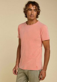 T-shirt à manches courtes, col rond en coton rose clair, avec une texture subtile et un petit logo près de l'ourlet.