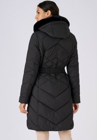 Zwarte gewatteerde jas met capuchon, voorzien van een ruitvormig doorgestikt patroon, elastische taille en zachte bontafwerking rond de kraag.