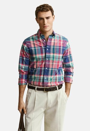 Junger Mann, der ein bunt kariertes Hemd mit hochgekrempelten Ärmeln, cremefarbene Hose und einen geflochtenen dunklen Gürtel trägt, hält einen braunen Taschengurt.