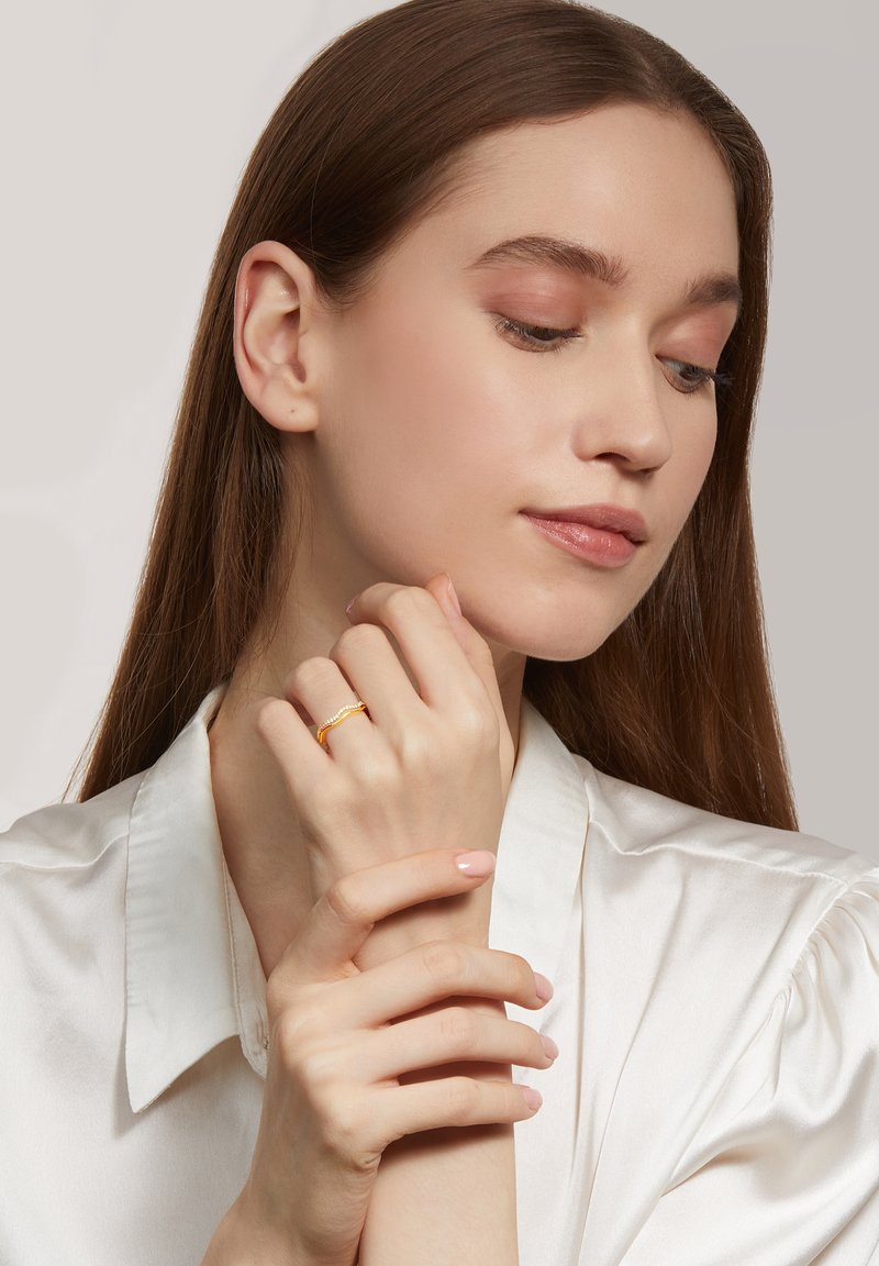 Gold ring with a twisted design worn on a hand, featuring smooth skin and a light pink nail polish against a neutral background.