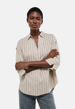 Femme aux cheveux courts portant une chemise beige à rayures à manches longues avec un col et un jean bleu, debout les bras croisés.